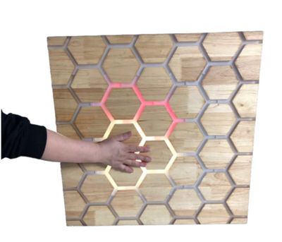 A hand touching a wooden wall mounted honeycomb interactive panel with LED backlighting. 