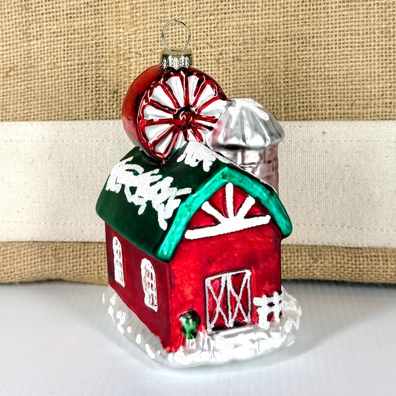 Glass red barn Christmas ornament with a red and white peppermint swirl topper and snow-dusted green roof.