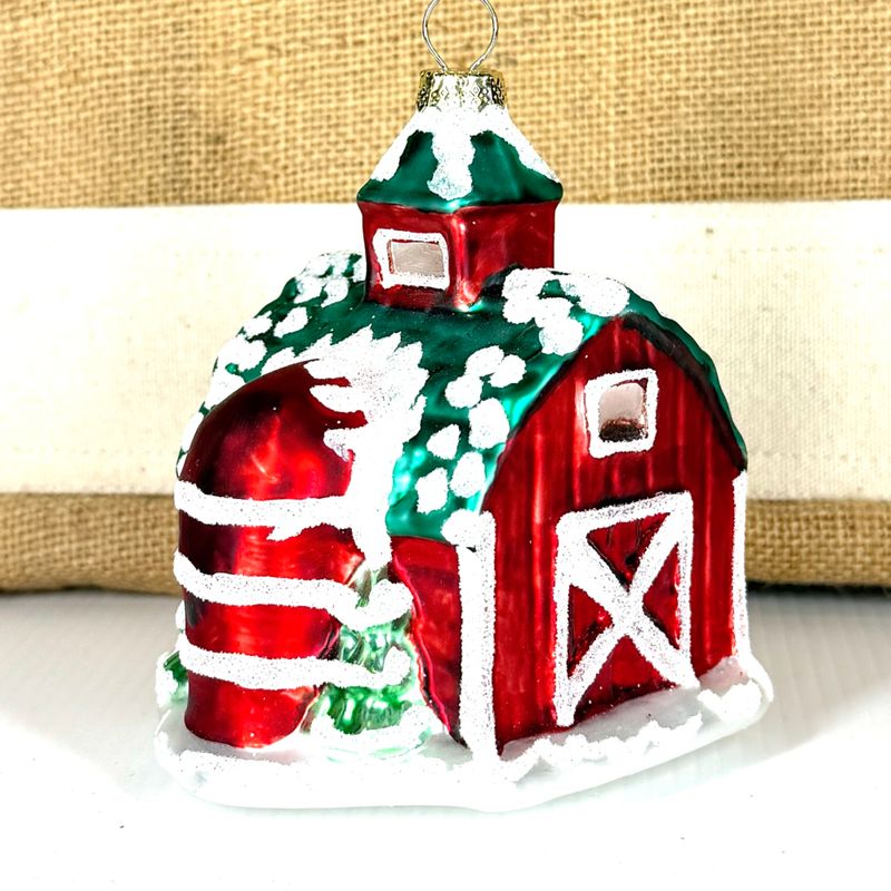 Side view of a glass barn ornament showing a red silo and frosted windows against a burlap background