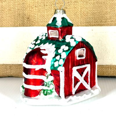 Side view of a glass barn ornament showing a red silo and frosted windows against a burlap background