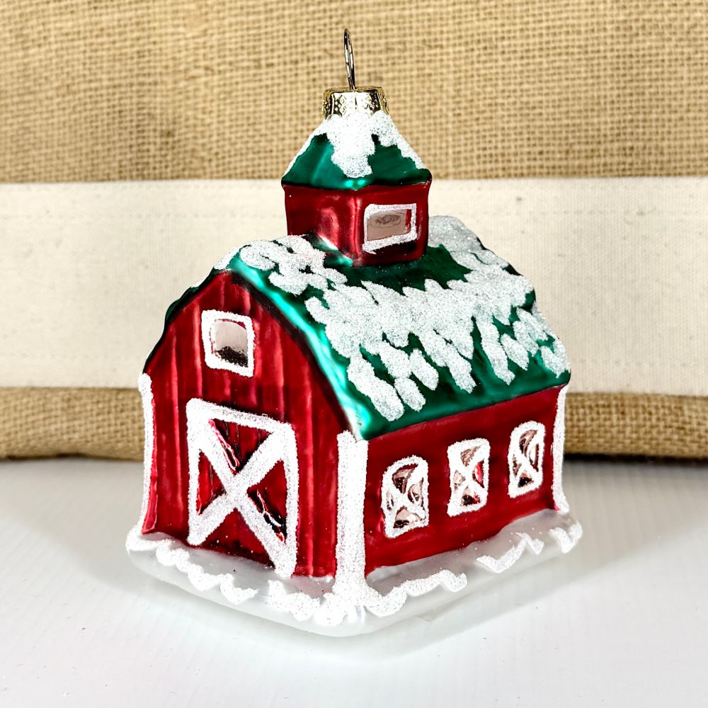 Front view of a metallic red barn glass ornament with snow-dusted green roof and white barn doors