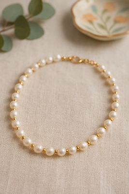 Kette aus weißen und goldfarbenen Perlen liegt auf einer beigen Tischdecke aus Leinen. 