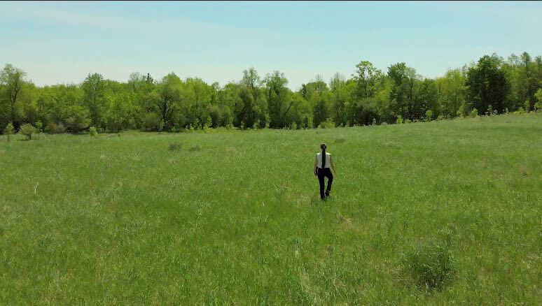 Young Woman walks straight away in Grassy Field while carrying an office book. (2) Young Woman walks straight away in Grassy Field while carrying an office book. (2)