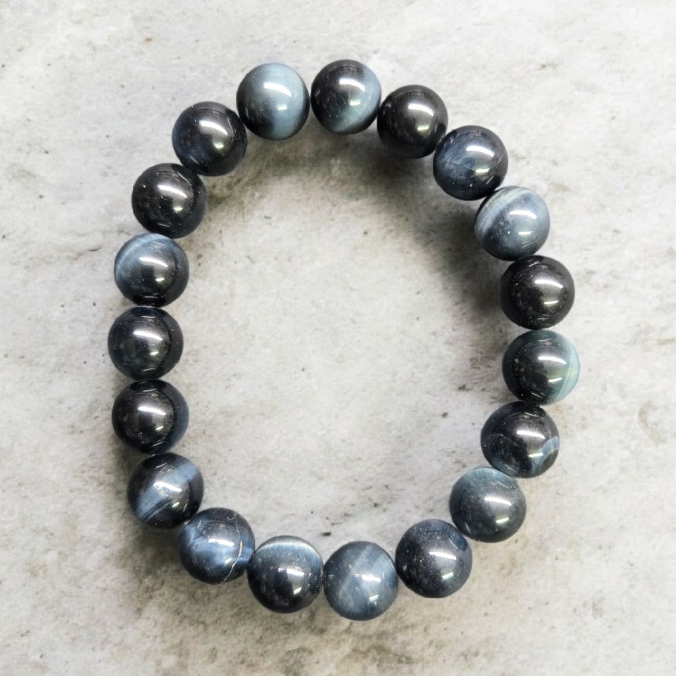 BLUE TIGER EYE 10MM BRACELET Blue Tiger Eye 10mm stretch crystal bracelet with polished round beads on elastic cord displayed on a light stone background.