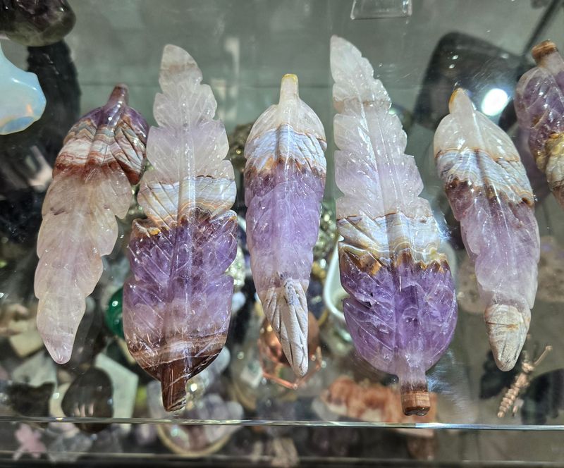 Amethyst Lace Agate feather crystal carvings with purple, cream, and banded patterns displayed together in a glass case