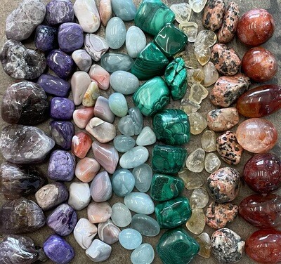 Assorted healing crystals and tumbled stones including amethyst, malachite, agate, citrine, and aquamarine arranged in rows.