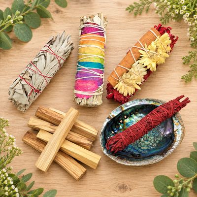Sage bundles, palo santo sticks, and a red smudge bundle in an abalone shell arranged on a wooden table with greenery.