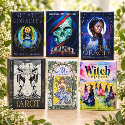 Six tarot and oracle decks displayed on a wooden table with soft greenery and white flowers in the background.