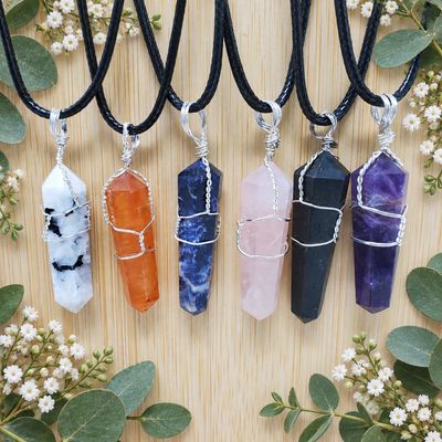 Six crystal point necklaces with silver wire wraps on a light wooden table framed by greenery and white flowers.