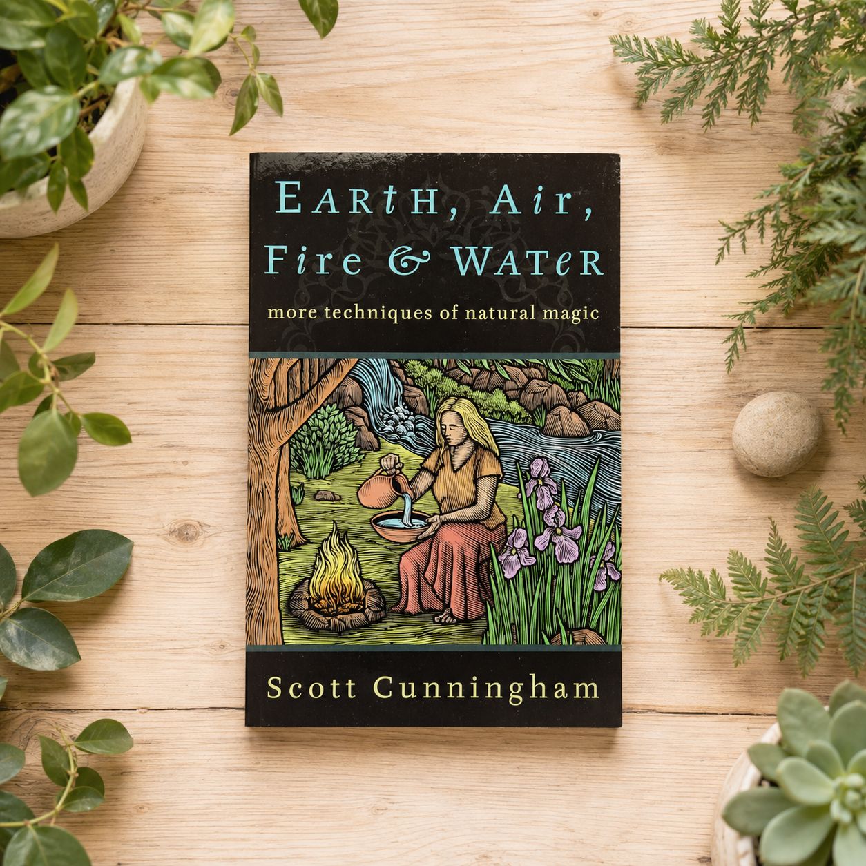 Earth, Air, Fire &amp; Water by Scott Cunningham on a light wooden table with greenery.