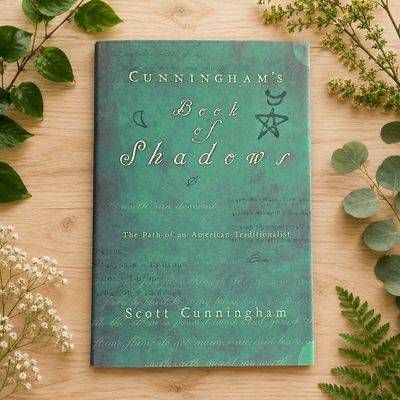 Cunningham’s Book of Shadows by Scott Cunningham on a light wooden table with greenery.