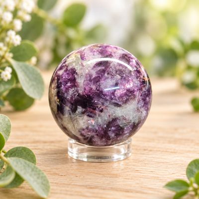 Small Lepidolite sphere with lavender, purple, and white natural patterning on a light wooden table with greenery.