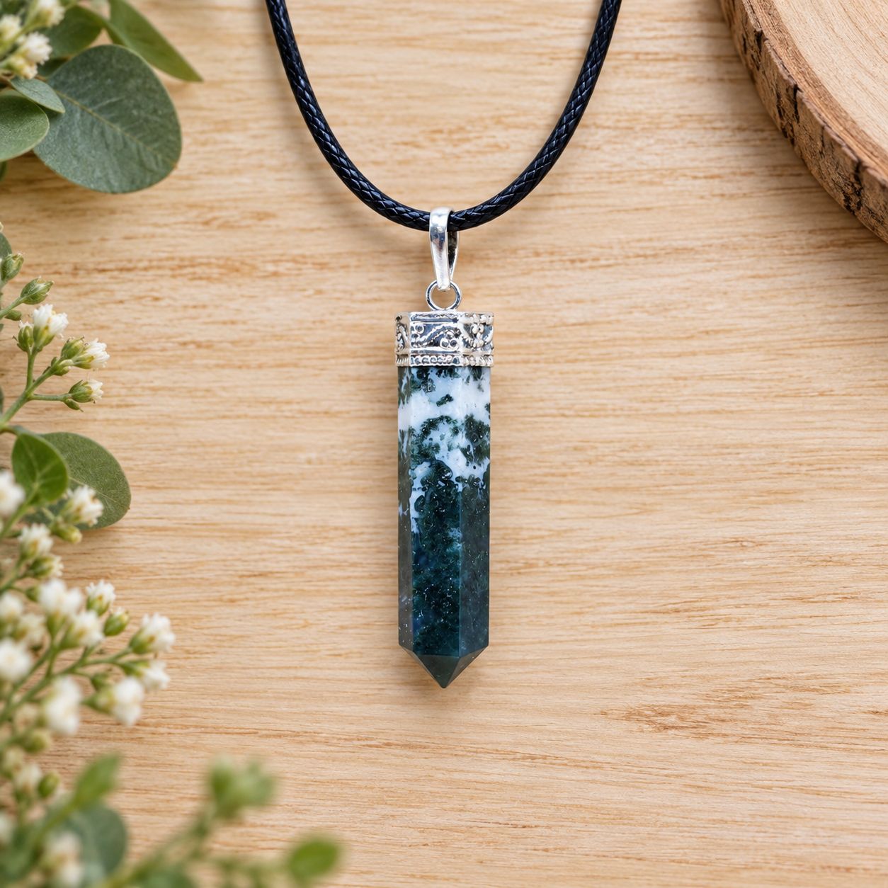 Moss agate pencil pendant necklace with silver tone cap on black cord displayed on light wood with greenery