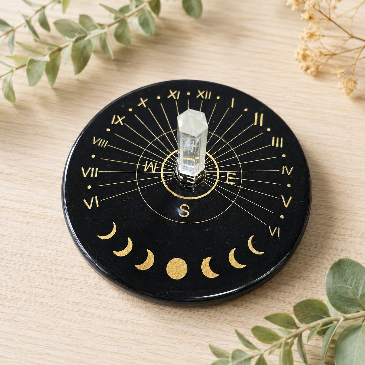 Black agate moon phase sun clock with clear quartz point on a light wooden table with greenery.