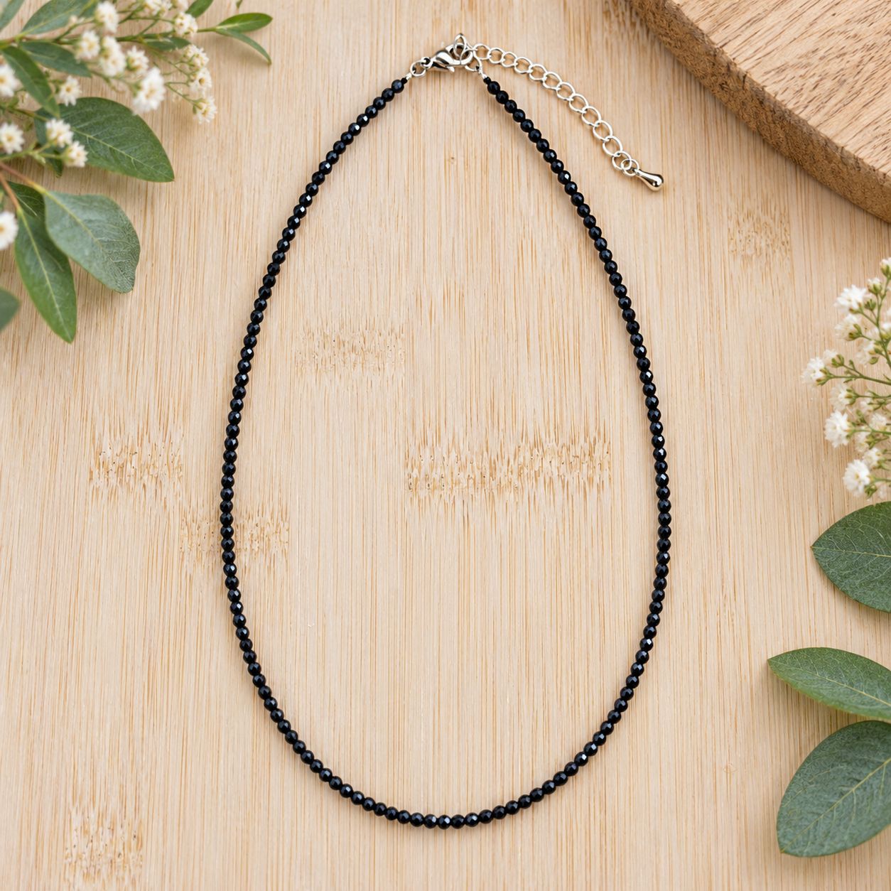 Black tourmaline 2mm faceted bead necklace with adjustable silver-tone clasp displayed on light wood with greenery.