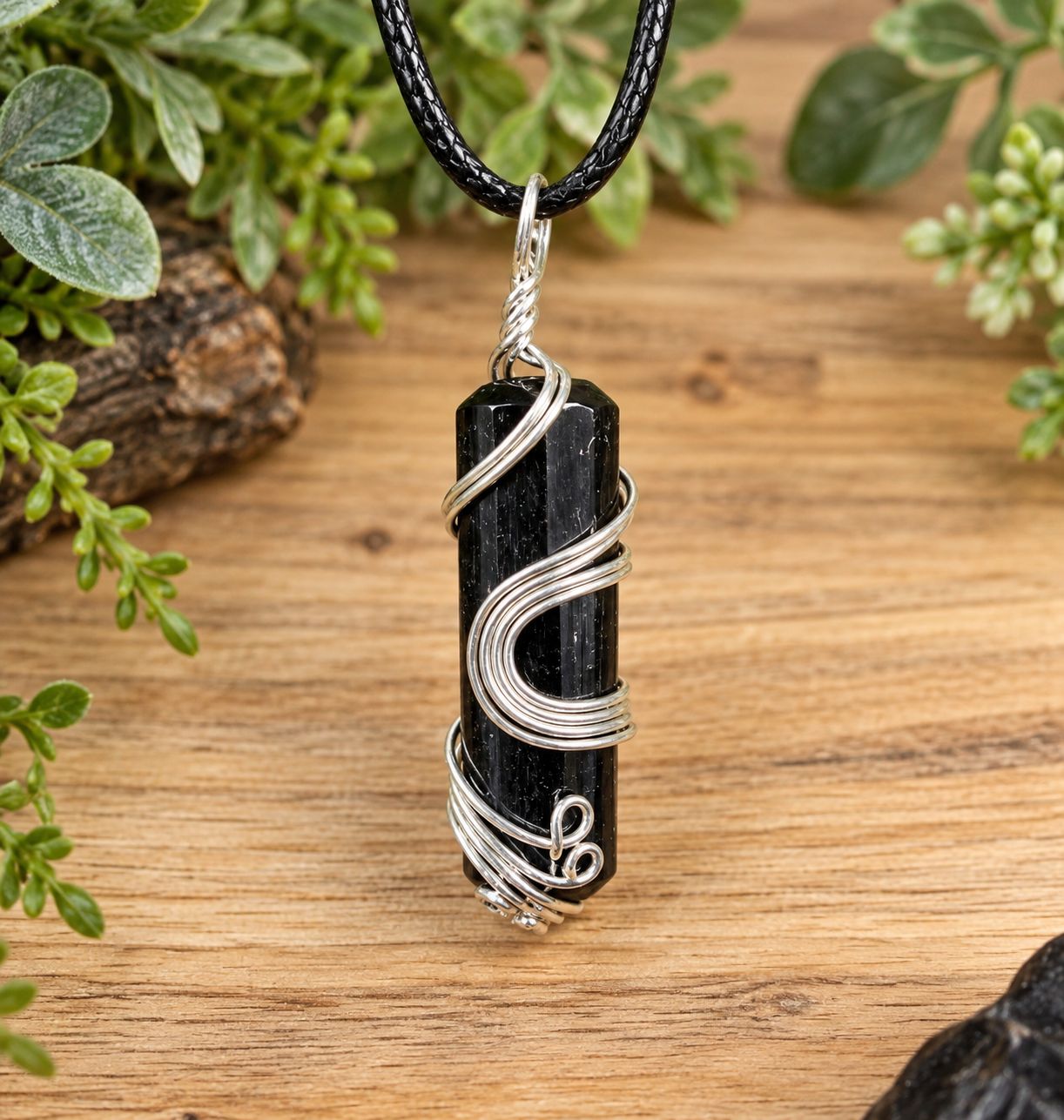 Ornate wire wrap black tourmaline necklace on black cord over light wood with greenery.