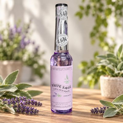 White Sage with Lavender Cologne bottle on a light wooden table with greenery, sage, and lavender.