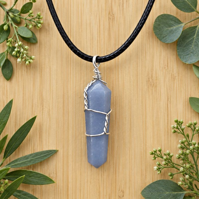 Angelite wire wrap pendant necklace on black cord over light wood with greenery.