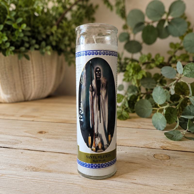 White Santa Muerte prayer jar candle on light wooden altar table with greenery background at Arkadys Metaphysical Gifts.