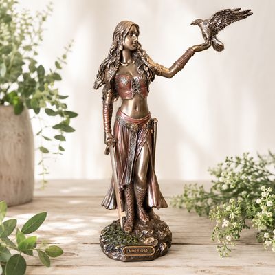 Hand painted Morrigan Celtic goddess statue with raven, sword, bronzed finish, and rocky base on a light wooden table
