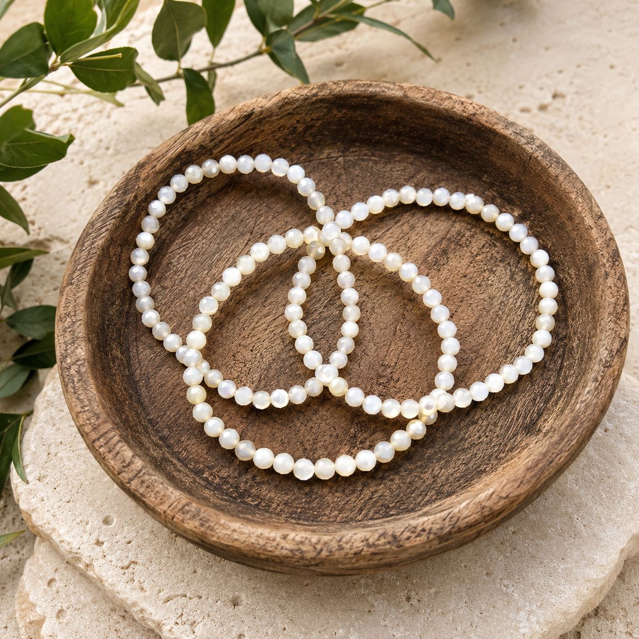 4mm Mother of Pearl faceted bracelets in rustic wooden bowl on stone altar with greenery.