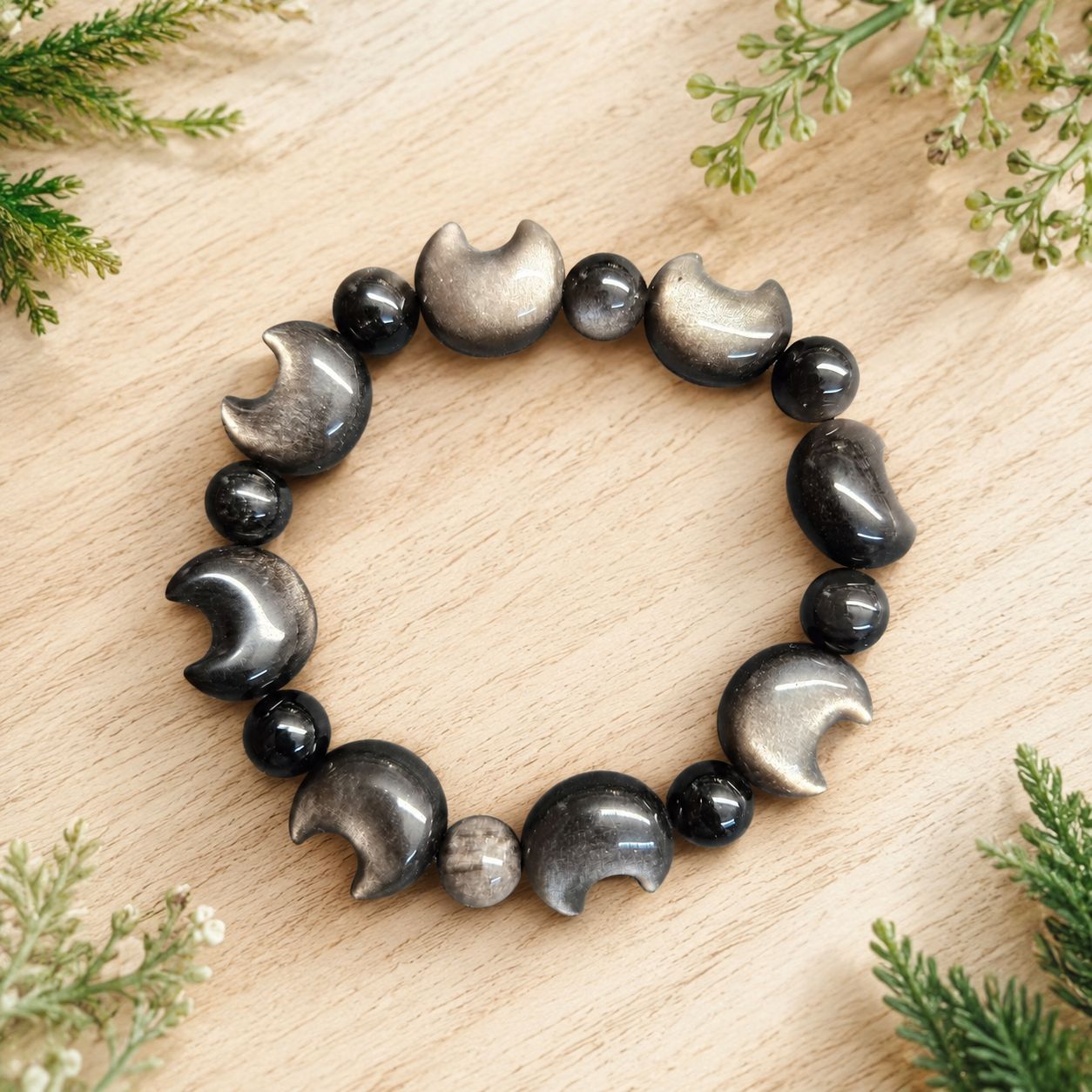 SILVER SHEEN OBSIDIAN MOON BRACELET Silver sheen obsidian moon bracelet on light wooden table with greenery