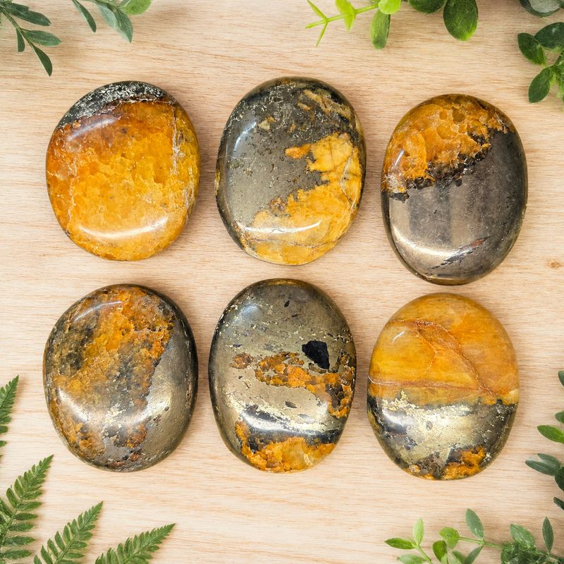 Golden Healer and Pyrite plump palm stones on a light wooden table with greenery