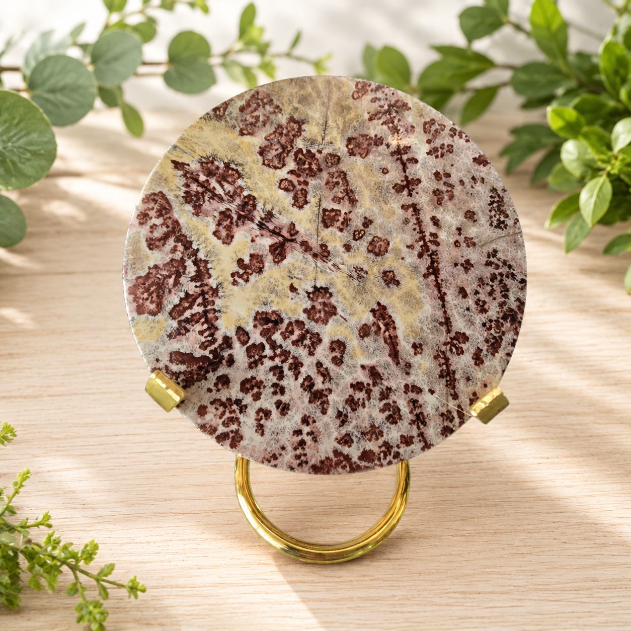 Picture jasper disc slab displayed on a gold stand on a light wooden table with greenery.
