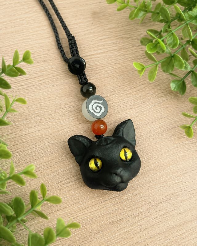 Obsidian cat head bag charm with yellow eyes and decorative beads on a light wooden table with greenery.