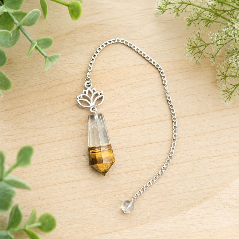 Tiger eye and quartz lotus pendulum on light wood with greenery.