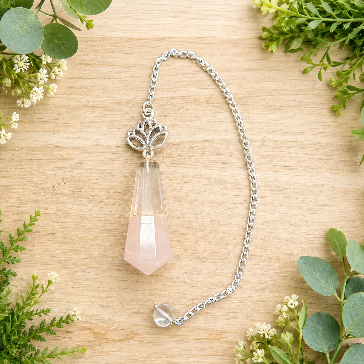 Rose quartz and quartz lotus pendulum on light wood with greenery.