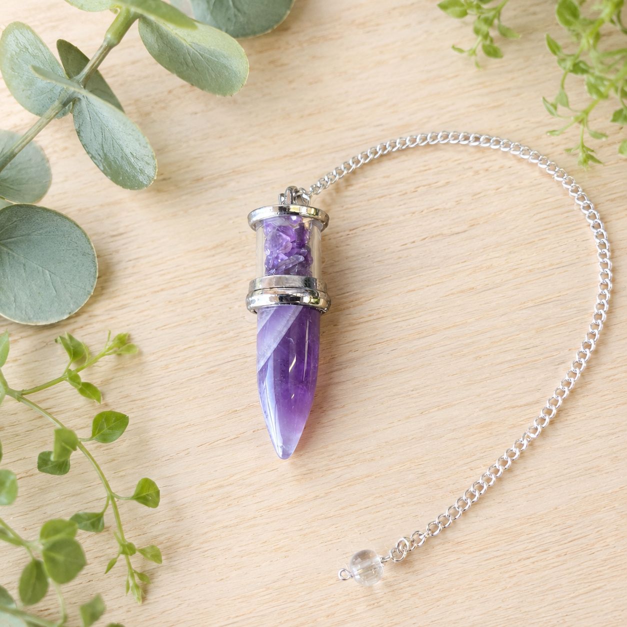 Amethyst Bullet Chips Pendulum with silver-toned chain on a light wooden table with greenery
