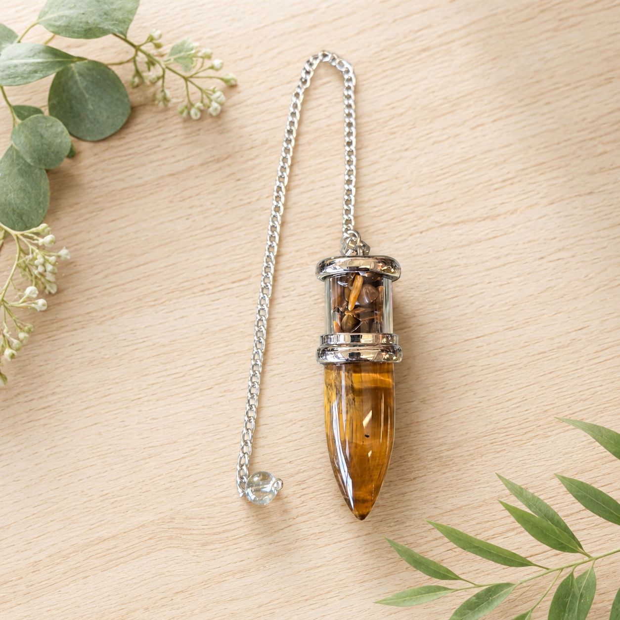 Tiger Eye Bullet Chips Pendulum with silver-toned chain on a light wooden table with greenery