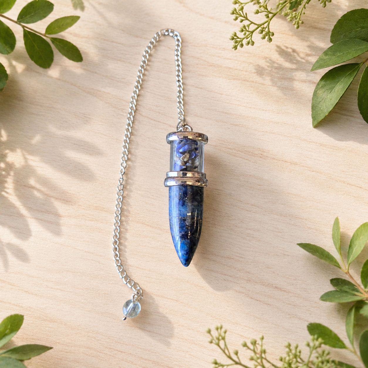 Lapis Lazuli Bullet Chips Pendulum with silver-toned chain on a light wooden table with greenery