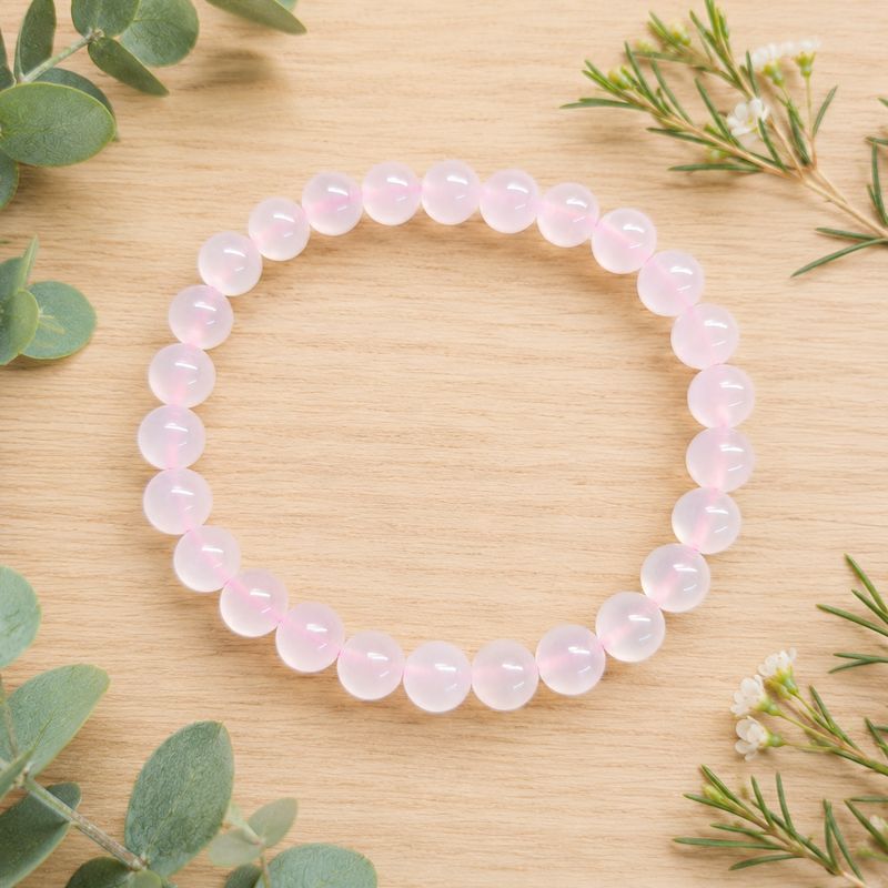 Pink calcite 8mm gemstone bracelet displayed on a light wooden table and some greenery. 