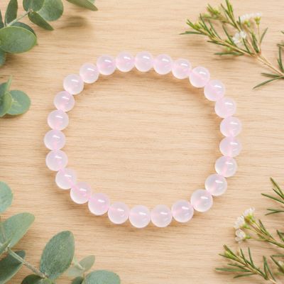 Pink calcite 8mm gemstone bracelet displayed on a light wooden table and some greenery. 