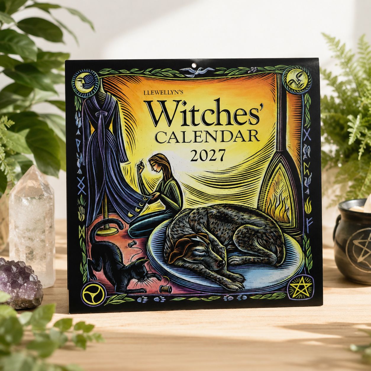 Front cover of Llewellyn’s 2027 Witches’ Calendar displayed on a light wooden table with greenery.