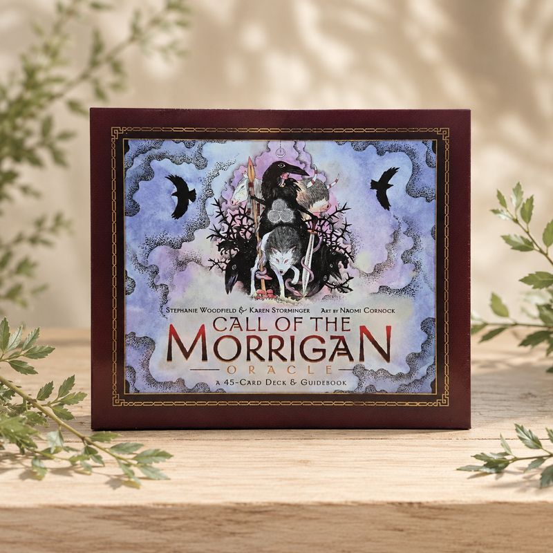 Front of Call of the Morrigan Oracle 45-card deck and guidebook displayed on a light wooden table with greenery