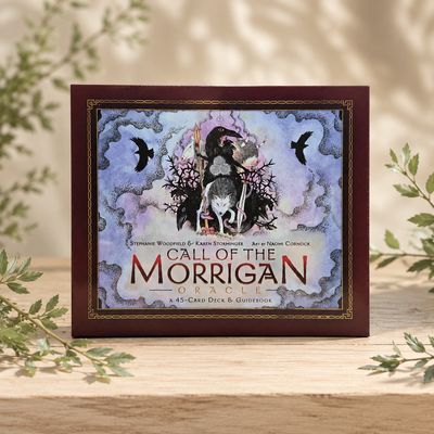 Front of Call of the Morrigan Oracle 45-card deck and guidebook displayed on a light wooden table with greenery