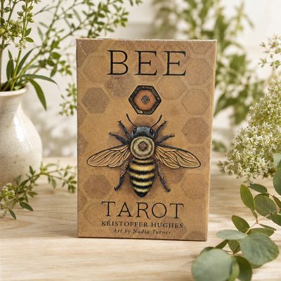 Bee Tarot deck by Kristoffer Hughes with honeybee artwork displayed on a light wooden table with greenery
