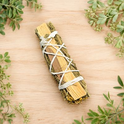 Rue and Palo Santo smudge stick displayed on a light wooden table with greenery