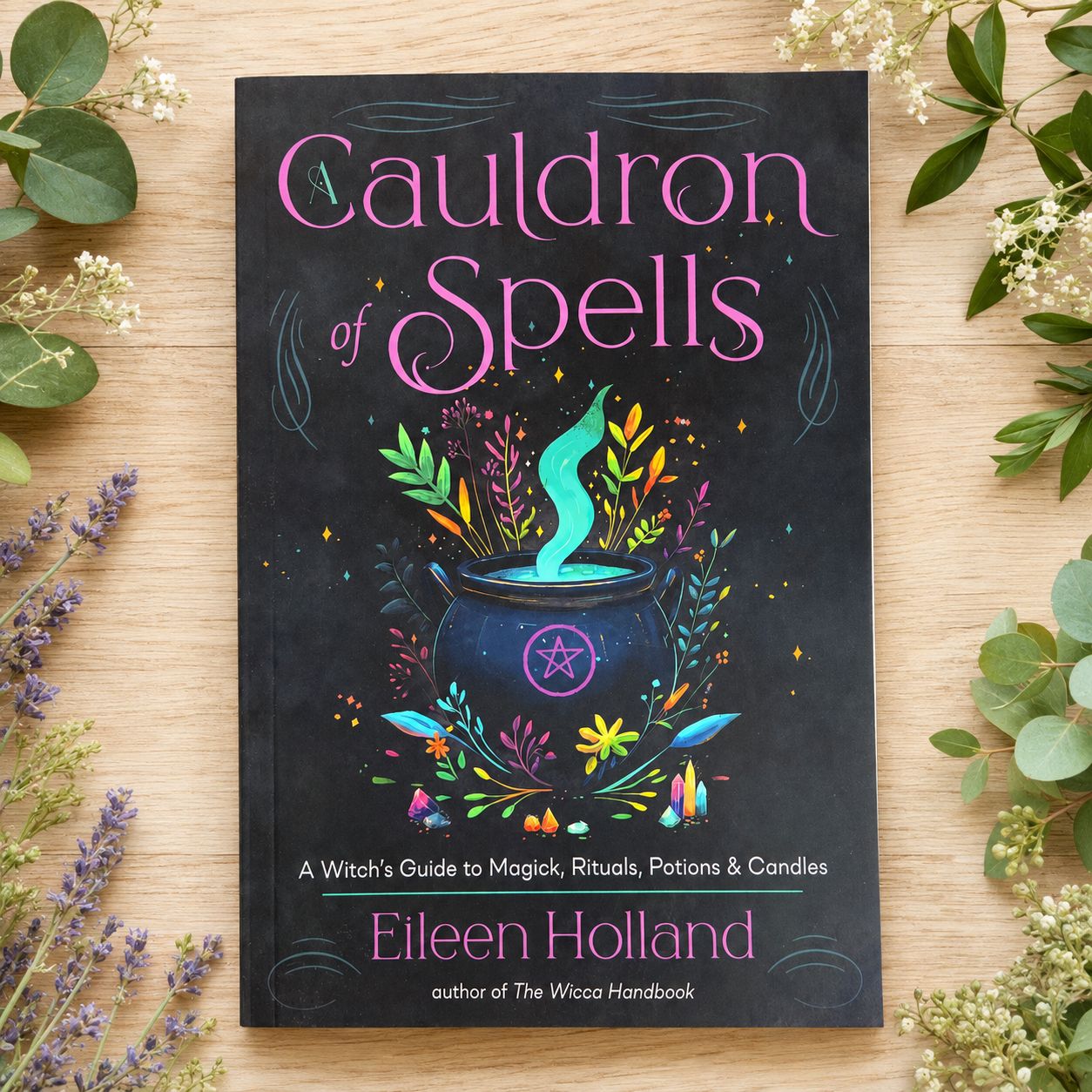 Cauldron of Spells by Eileen Holland displayed on a light wooden table surrounded by greenery.