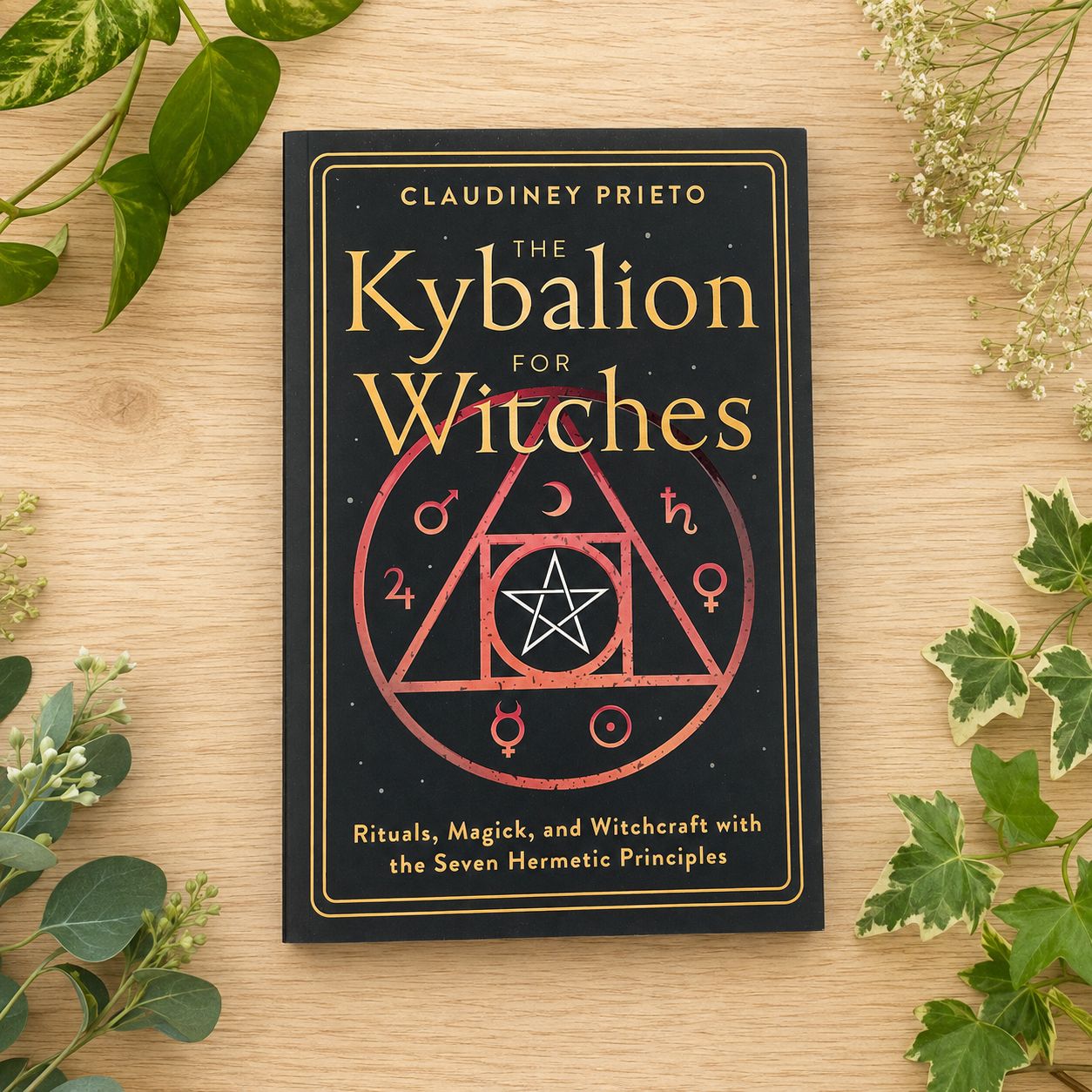 The Kybalion for Witches by Claudiney Prieto on a light wooden table with greenery.