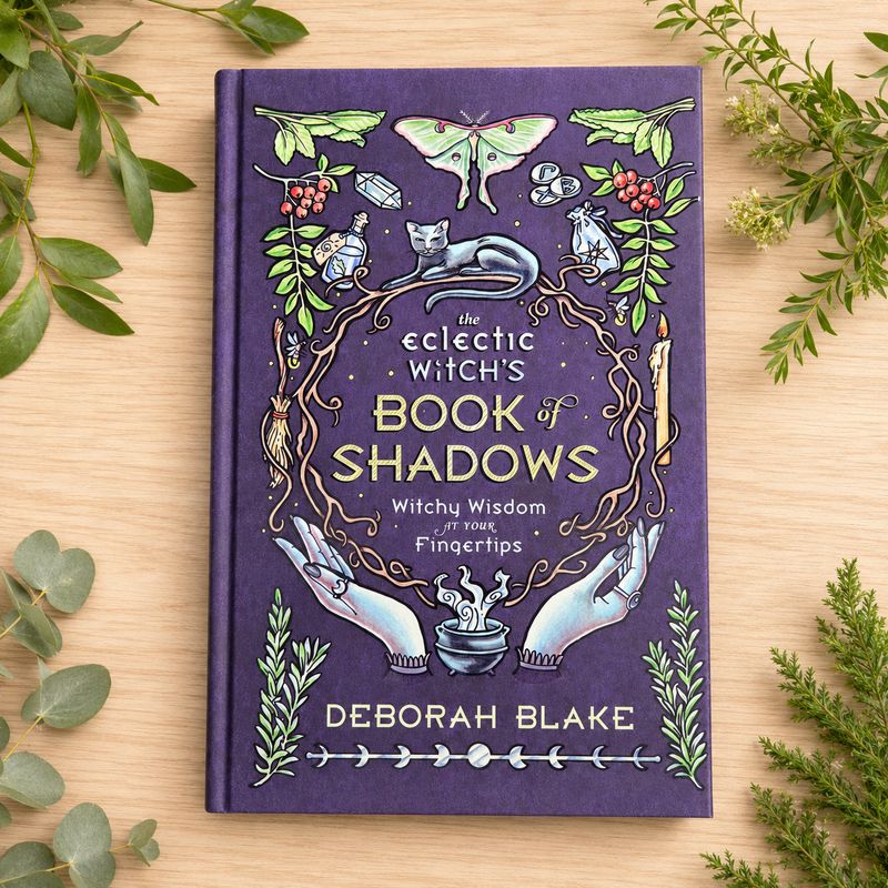 The Eclectic Witch’s Book of Shadows by Deborah Blake on a light wooden table with greenery.
