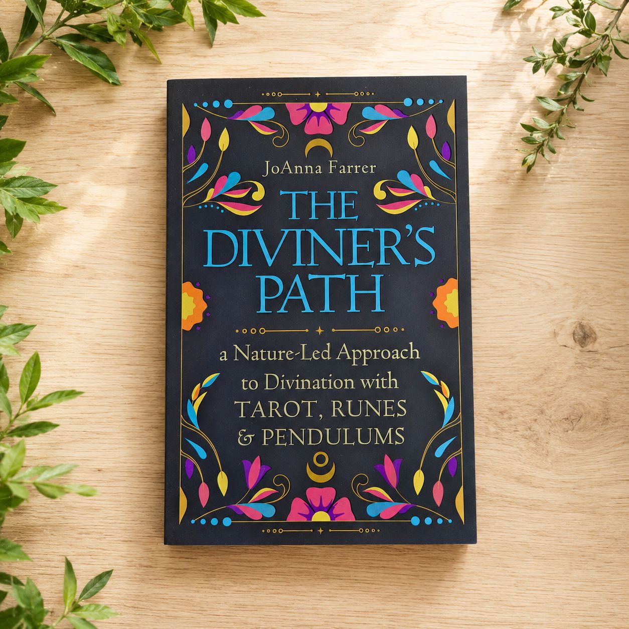 The Diviner’s Path by JoAnna Farrer displayed on a light wooden table with greenery.