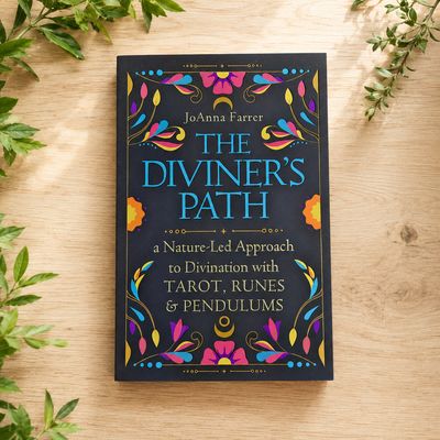 The Diviner’s Path by JoAnna Farrer displayed on a light wooden table with greenery.
