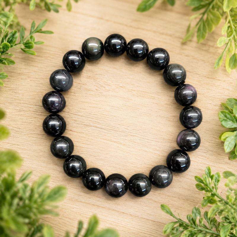 Rainbow Obsidian 10mm gemstone bracelet on a light wooden table with greenery.