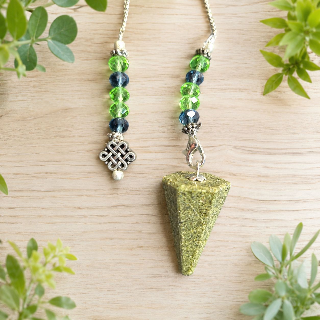 Close-up of an Epidote Celtic knot pendulum with green and blue beads on a light wooden table