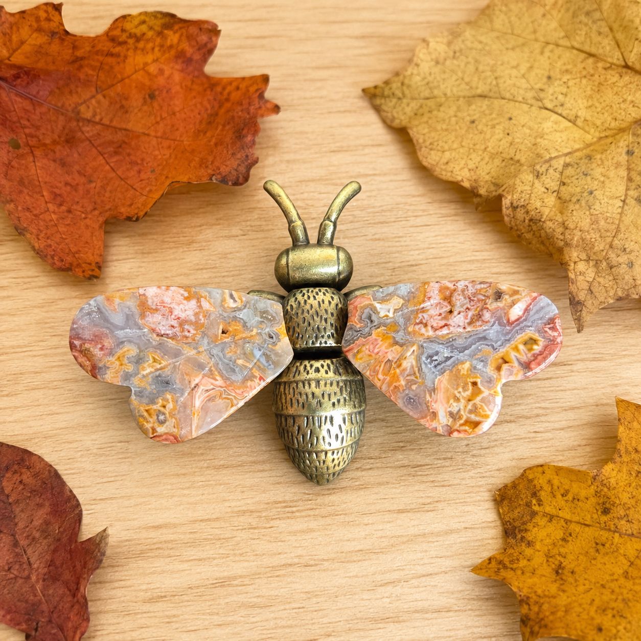 Crazy Lace Agate metal bee with gemstone wings displayed on a light wooden table with fall leaves.