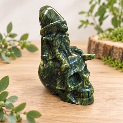 Side one of Nephrite jade gnome sitting on skull carving displayed on a light wooden table with greenery.